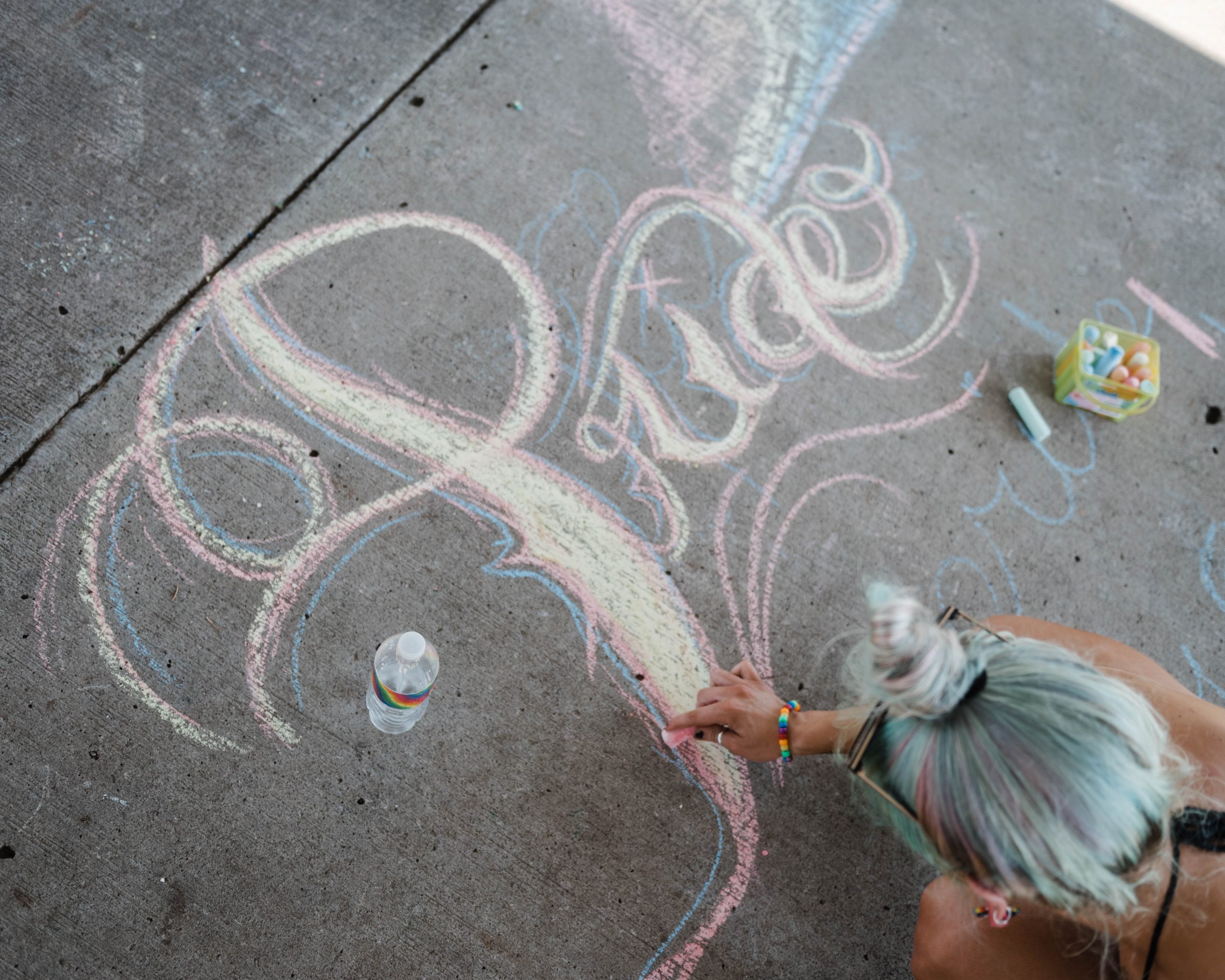 A person with light blue hair (seen from above and behind) draws the word "Pride" in calligraphy on a sidewalk