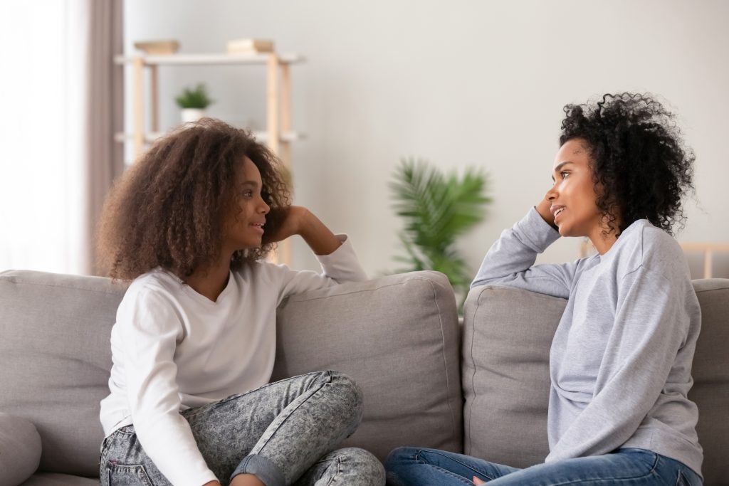 A Black woman casually talks with a Black teen while sitting on the couch