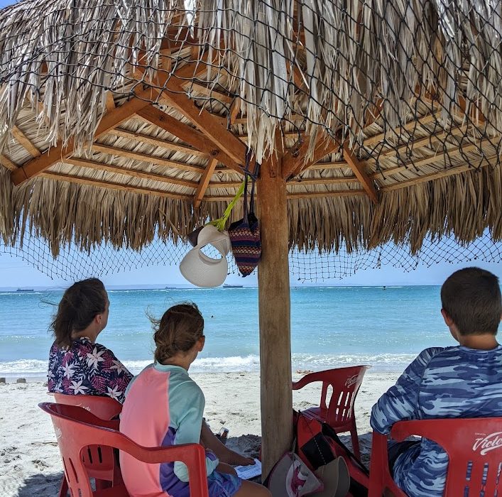 A woman and two kids sit on a beach under the shade of a thatch-roof palapa as they look out on the turquoise ocean.