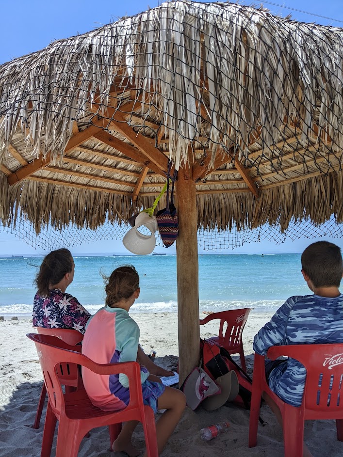 A woman and two kids sit on a beach under the shade of a thatch-roof palapa as they look out on the turquoise ocean.