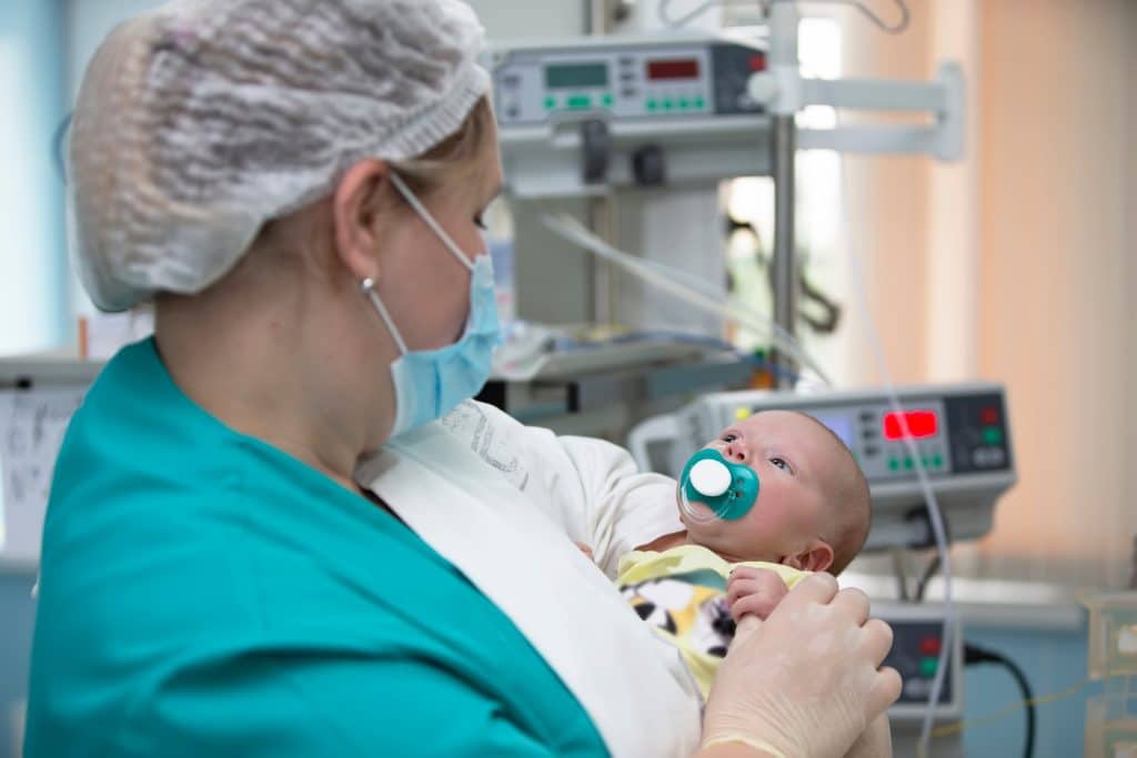 A nurse with a mask and hairnet on holds a baby in intensive care.