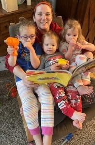 A woman sits in a chair with three young kids in her lap. They are all smiling at the camera