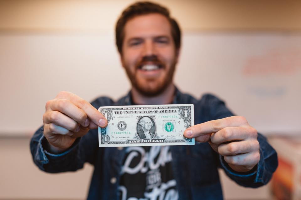 A man smiles while holding a dollar bill out in front of him