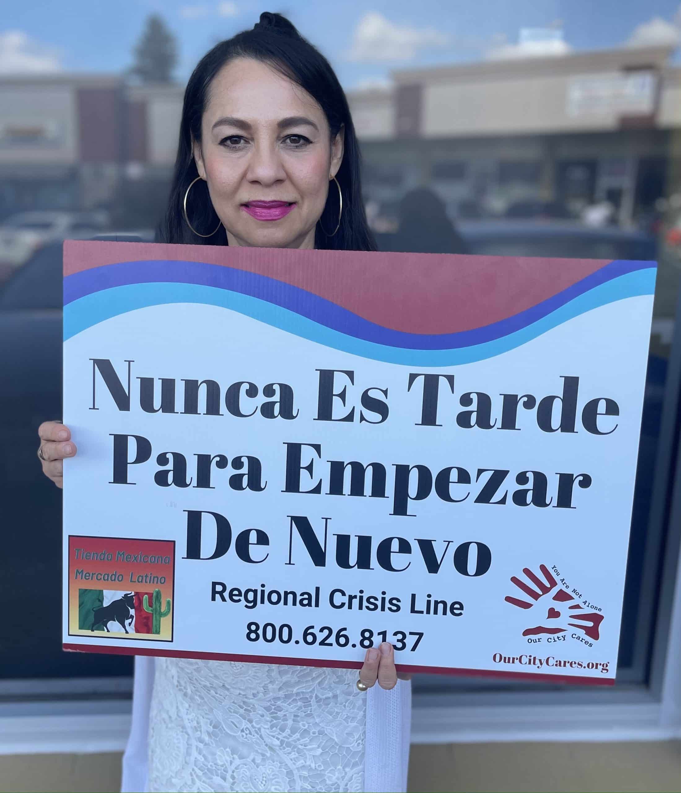 A woman with long black hair holds a sign that reads in Spanish, "Nunca Es Tarde Para Empezar De Nuevo" and in English, "Regional Crisis Line 800.626.8137