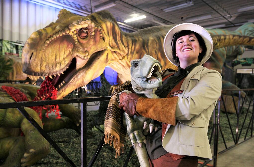 A woman in a safari costume holds a mini dinosaur figure in her arms, in front of a huge T-rex animatronic