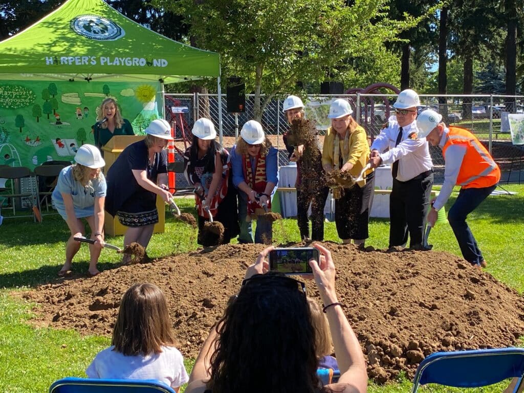 A group of people in hard hats plunge shovels into a pile of dirt for a playground groundbreaking ceremony