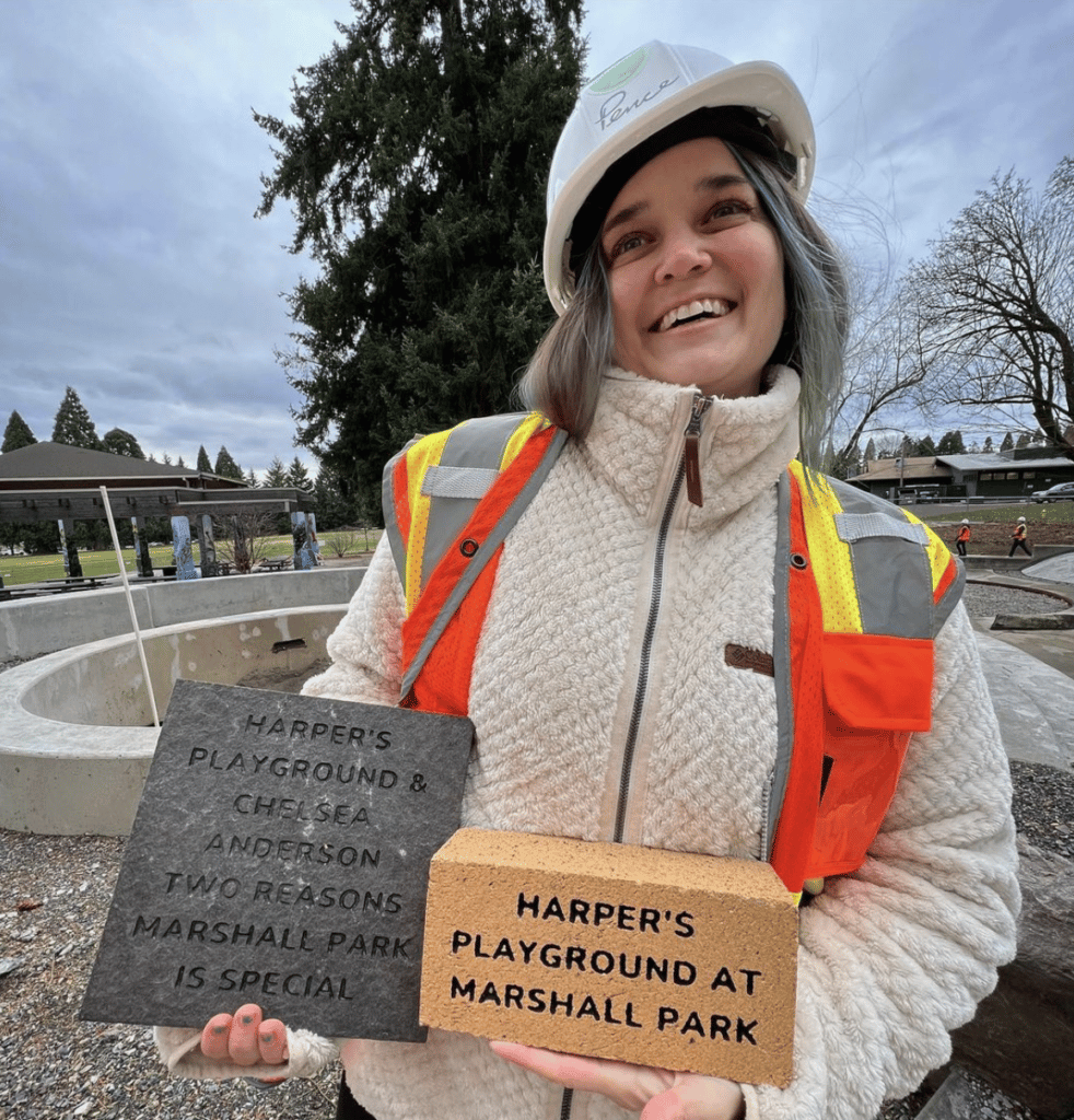 A woman in a hard hat and reflective vest smiles while holding a brick and a paver 