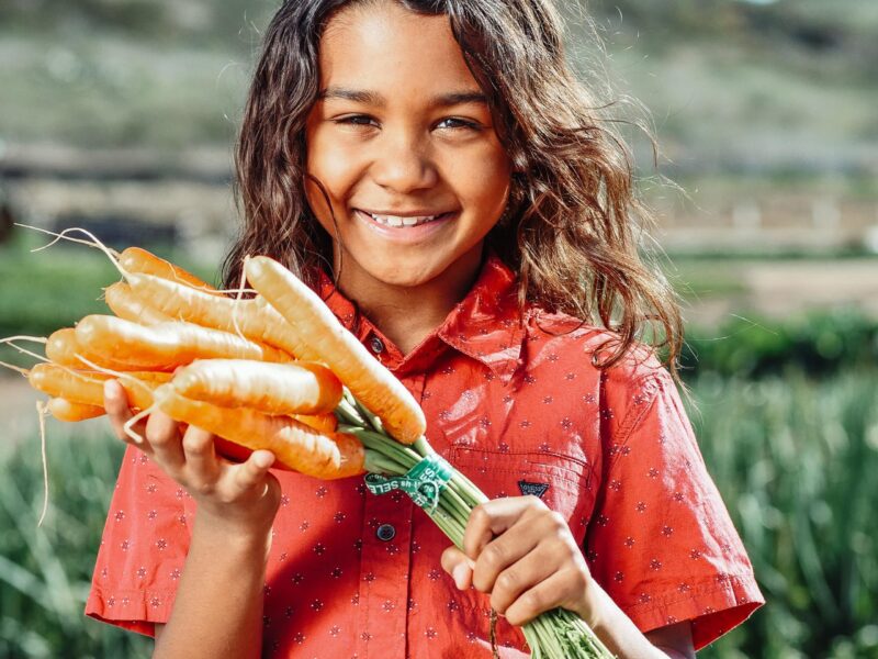 A child with mid-length curly brown hair and a red button-up shirt smiles while holding a bunch of carrots just pulled from the ground