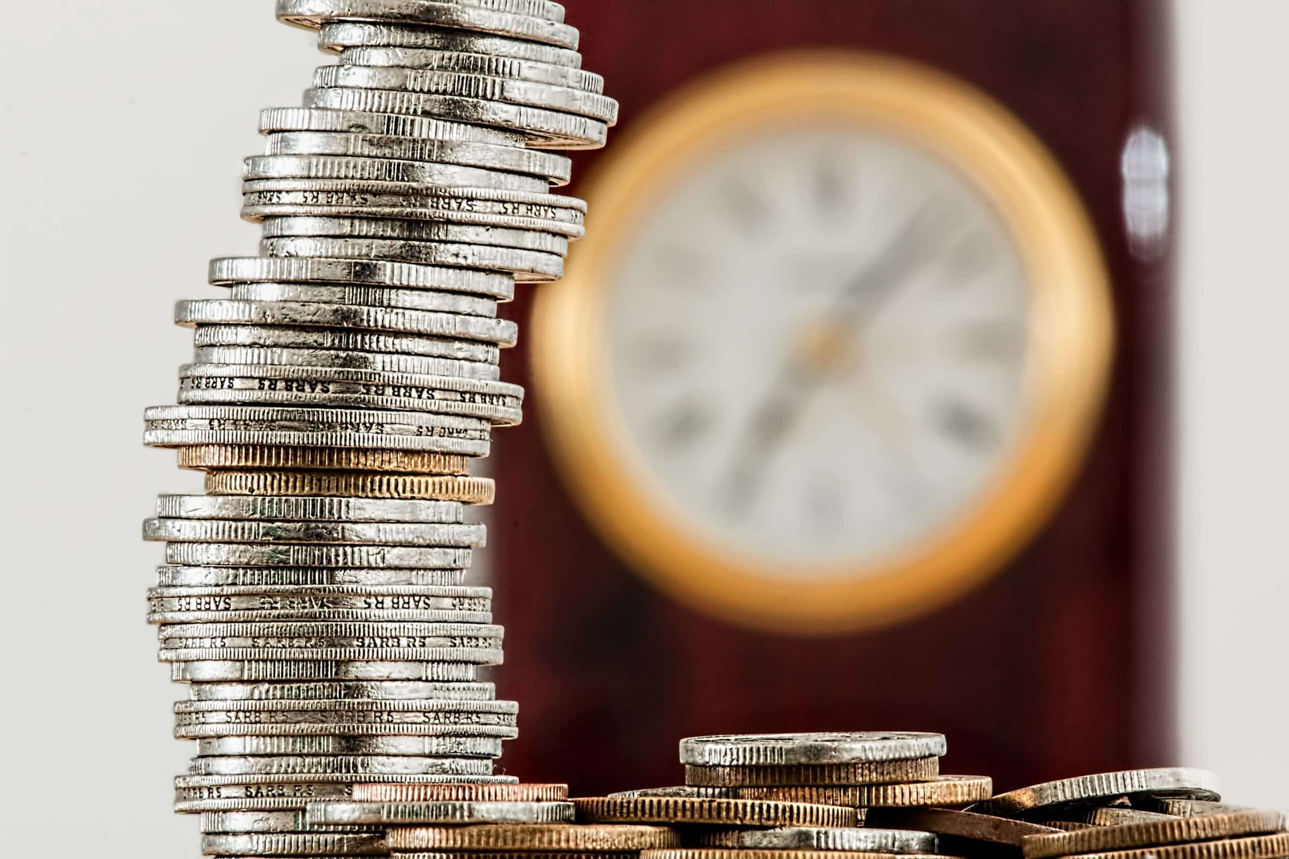 A stack of coins sits in front of a clock that is blurred in the background