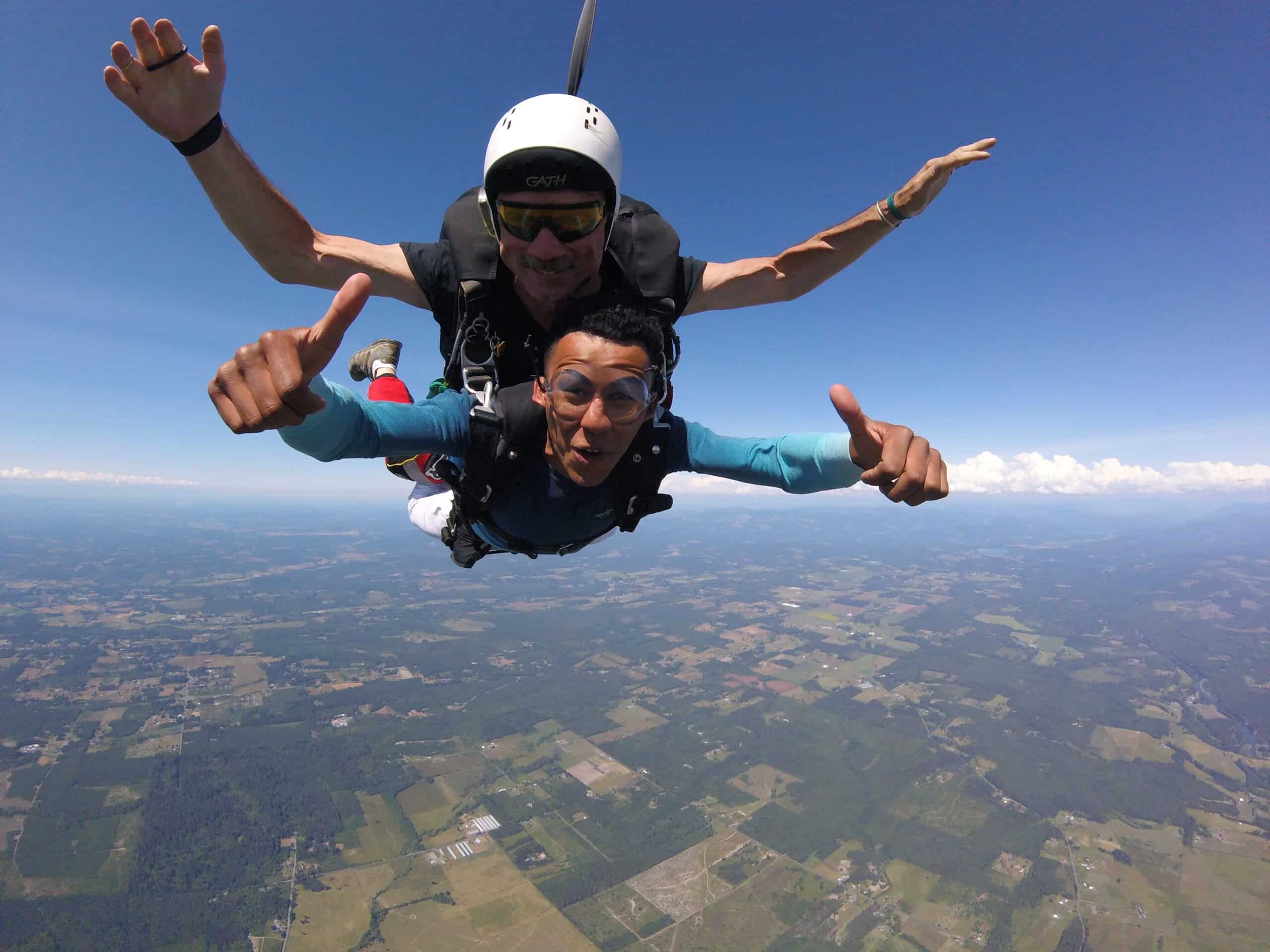 Two men are shown skydiving through the air with the earth thousands of feet below. Both are smiling at the camera and giving thumbs up.