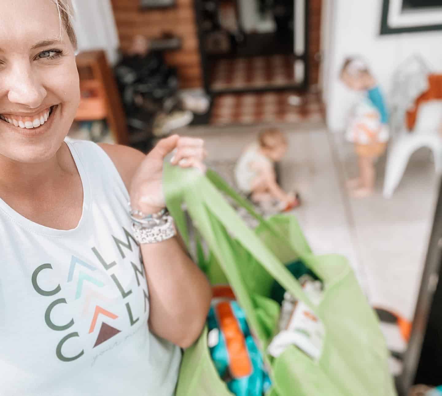 Brittany Clapp holds a canvas bag while smiling at the camera, with a couple of kids playing in the background