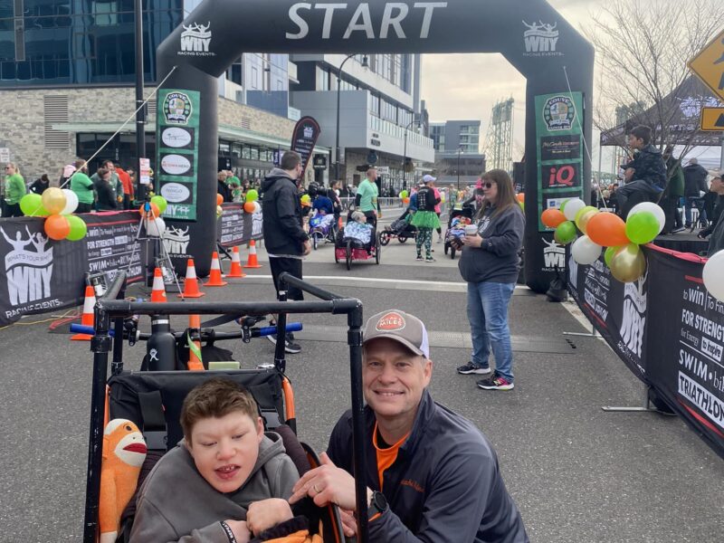 Micah Sn. sits in a wheelchair with his dad crouching next to him, smiling. A start line at a race is in the background.