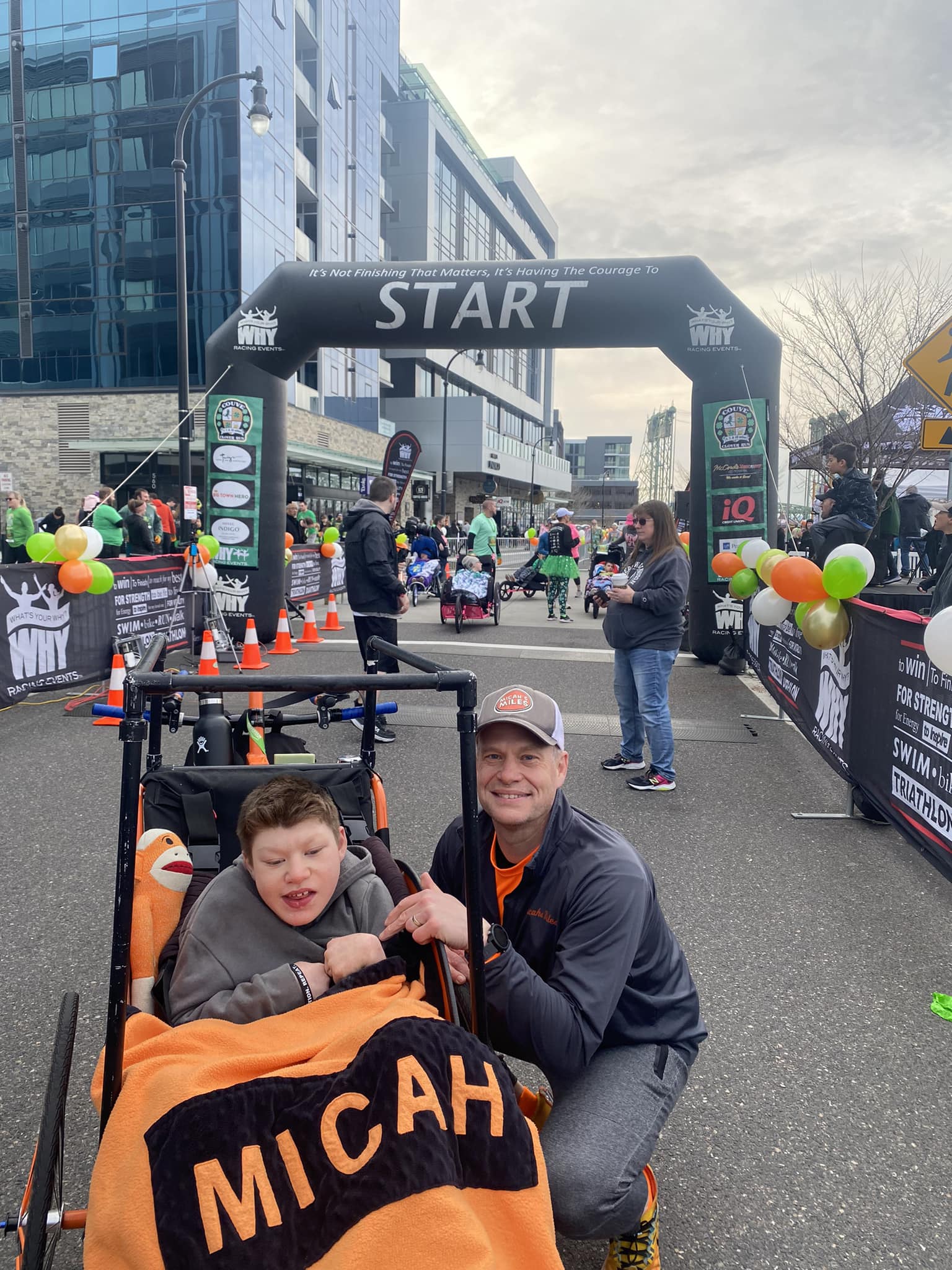 Micah Sn. sits in a wheelchair with his dad crouching next to him, smiling. A start line at a race is in the background.