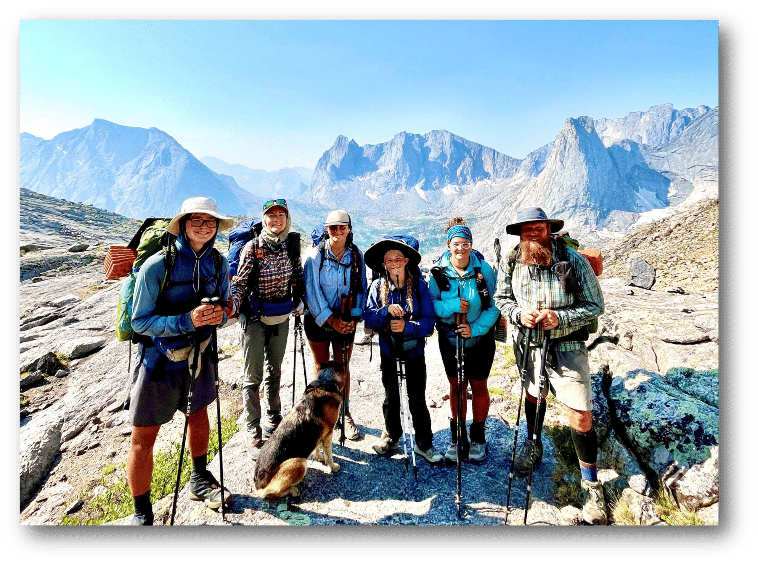 6 people and a dog smile on a mountain trail while wearing backpacking gear