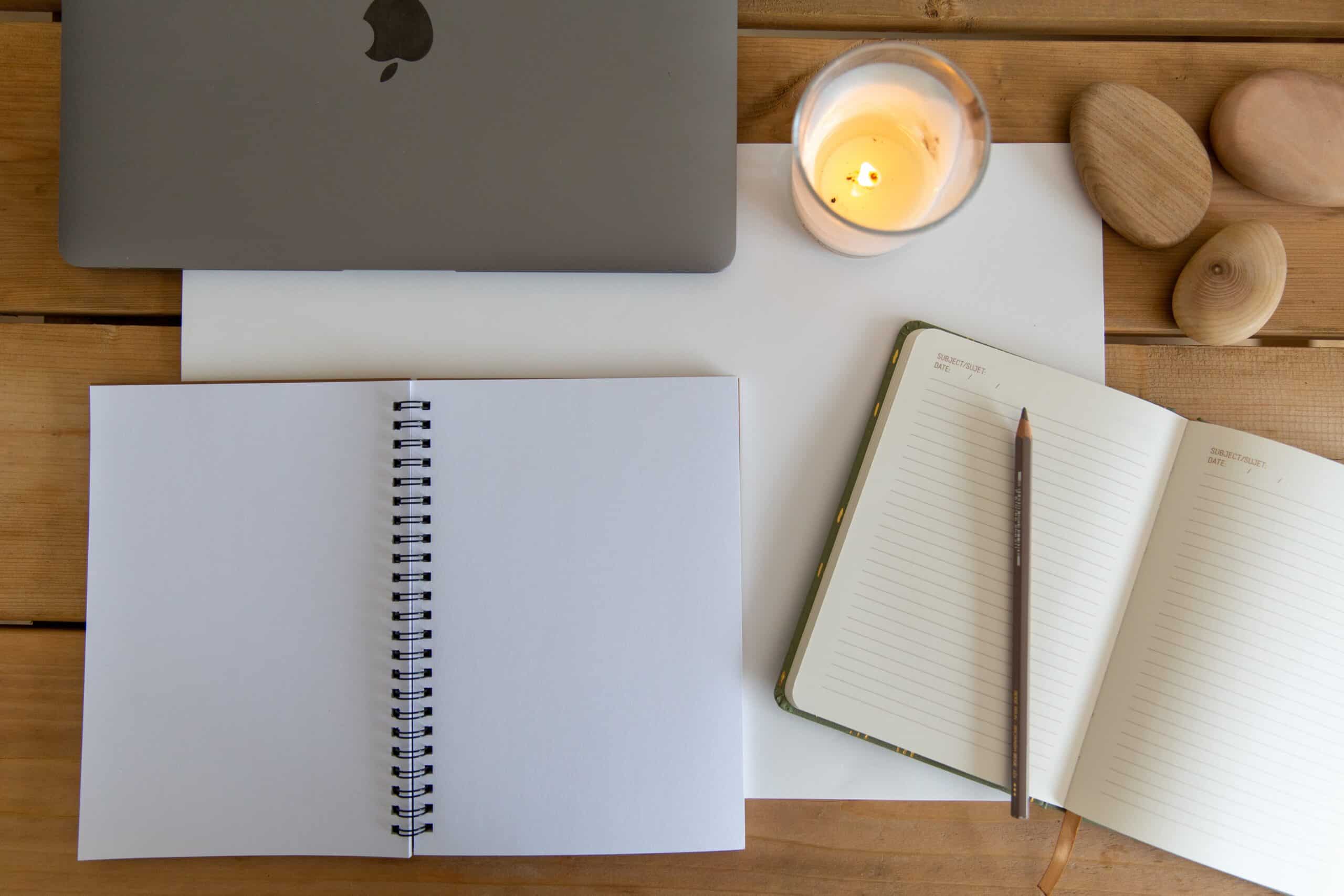 A blank notebook and journal site on a desk next to a laptop and a candle
