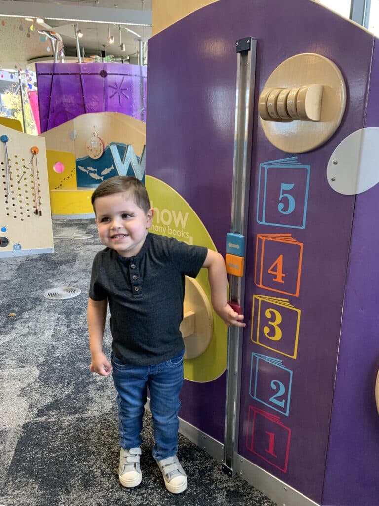 A young boy smiles in front of a colorful library display featuring numbers. Vancouver Community Library is on our list of free indoor things to do with kids in Vancouver and Clark County