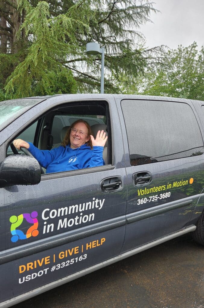 A Community in Motion volunteer smiles and waves from a gray van