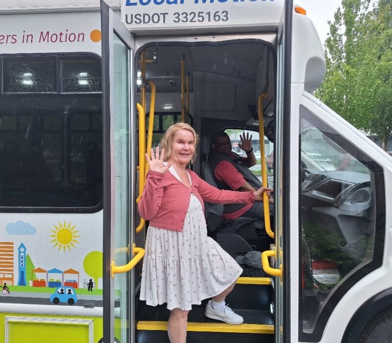 A woman waves from the steps of a small shuttle bus. The driver waves as well.