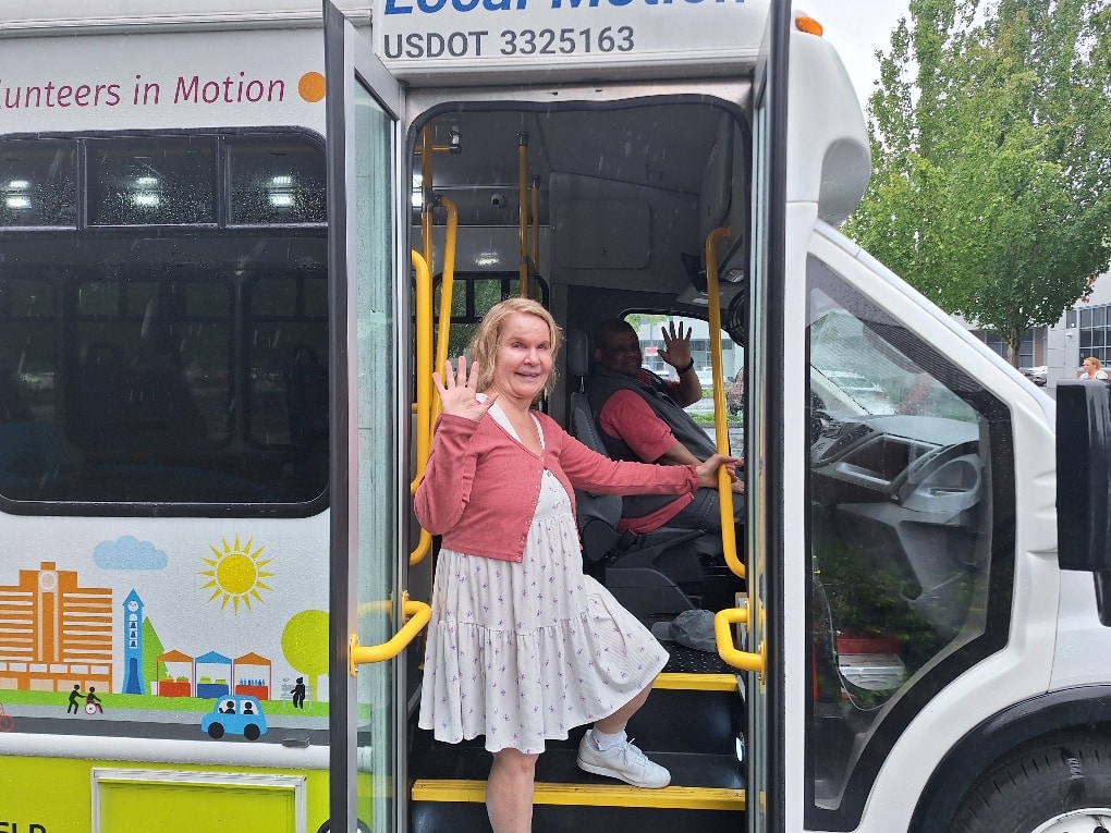 A woman waves from the steps of a small shuttle bus. The driver waves as well.