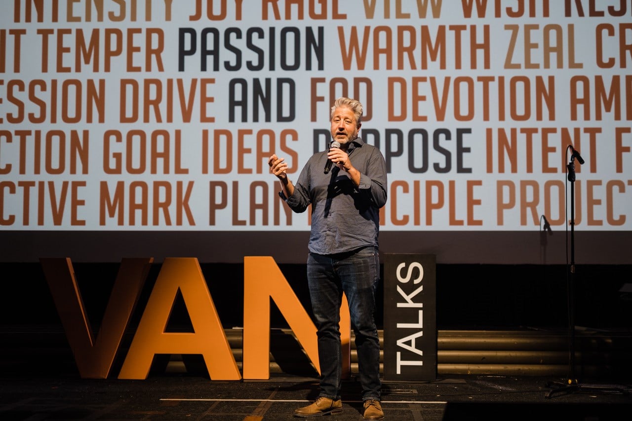 A man speaks on stage at Kiggins Theatre in Vancouver with a large sign reading VANTalks behind him.