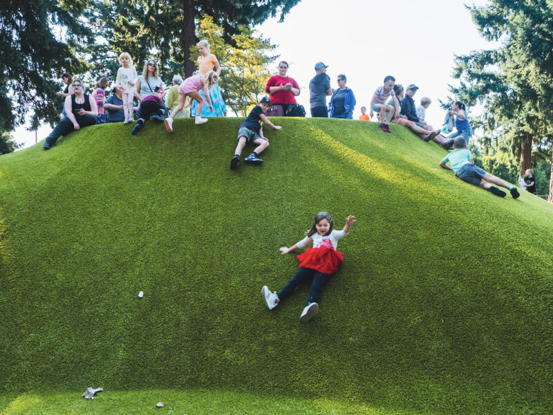A girl slides down a green hill with other kids at the top looking on