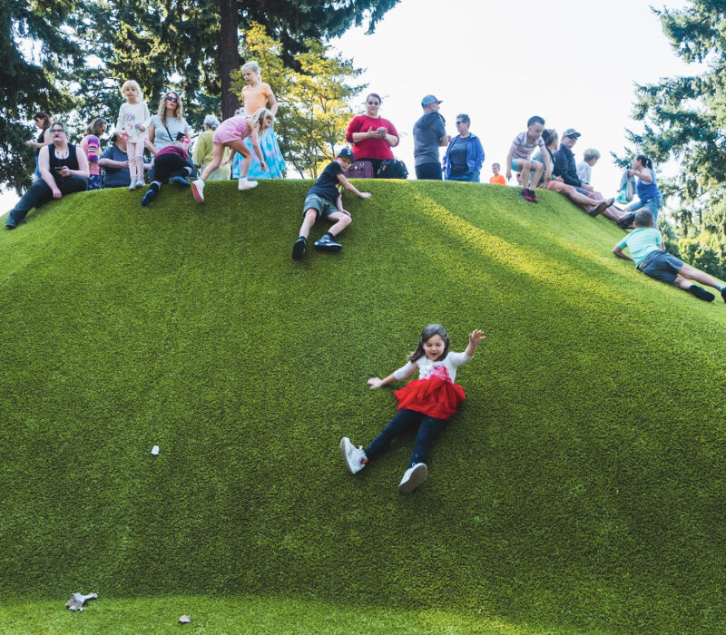 A girl slides down a green hill with other kids at the top looking on