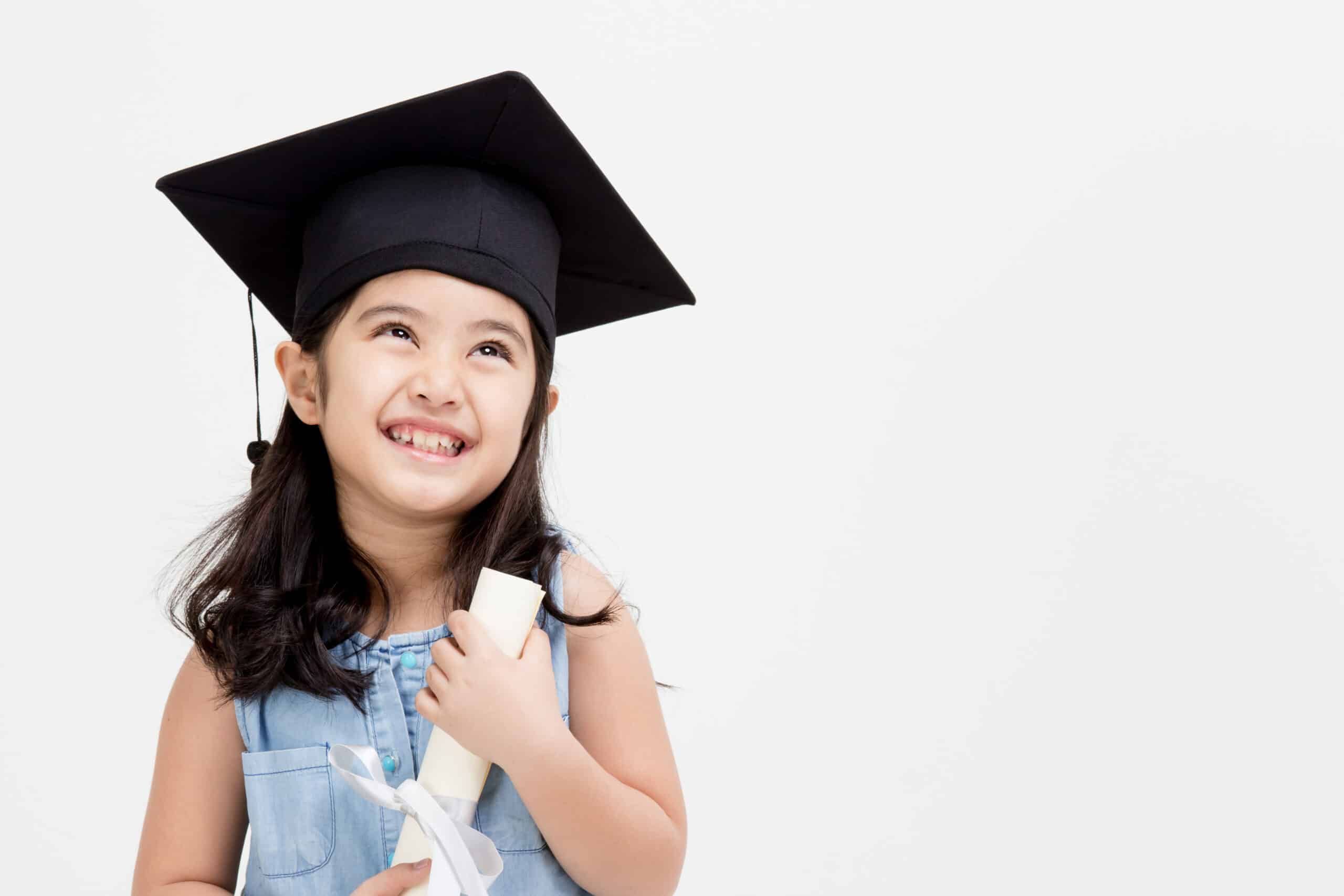 A girl with a black graduation cap holds a rolled up paper and smiles while looking dreamily up into the sky