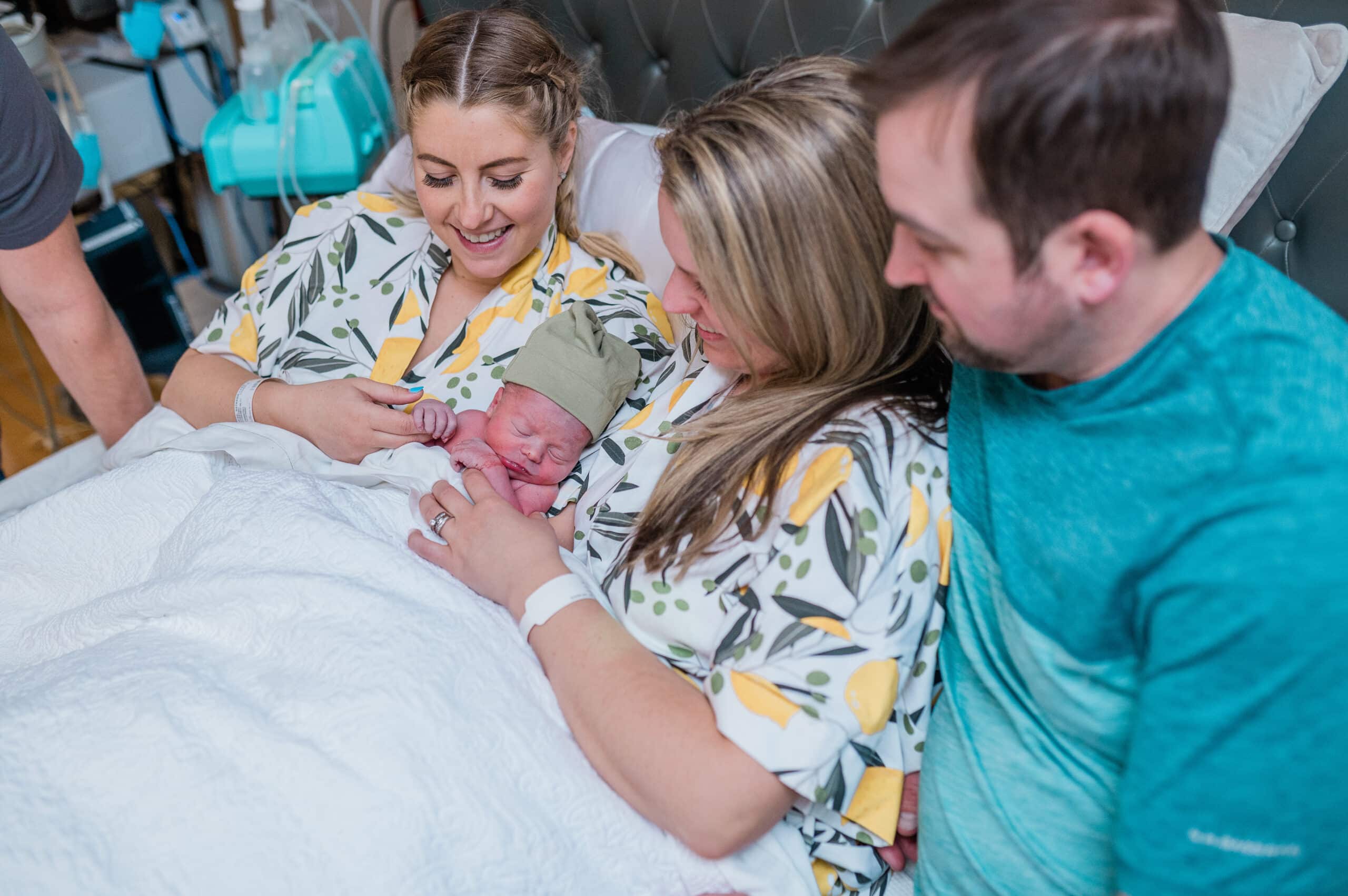 Two women in matching hospital gowns hold a baby between them