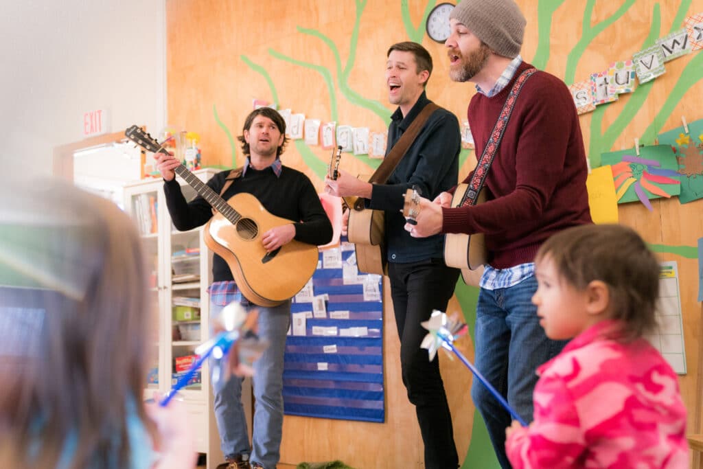 Three men sing while playing guitars in front of a group of children in a colorful room. The band Ants Ants Ants will perform as part of Wiggles and Giggles 2024