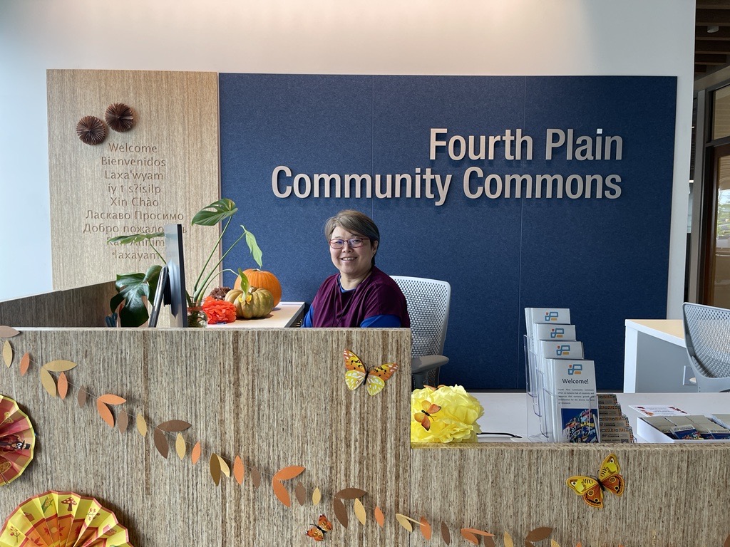 A Fourth Plain Forward staff member smiles behind the desk with a sign behind her reading "Fourth Plain Community Commons" 