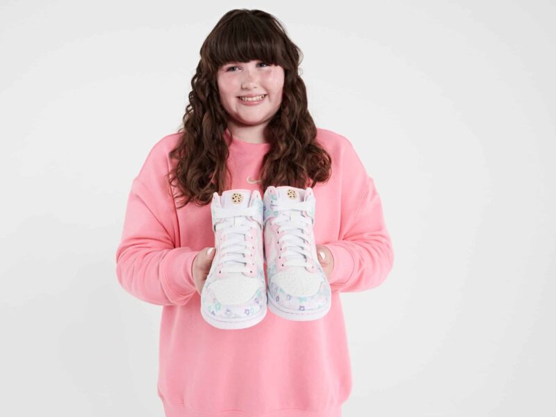 A teenage girl with long brown hair holds a pair of Nike shoes that she designed