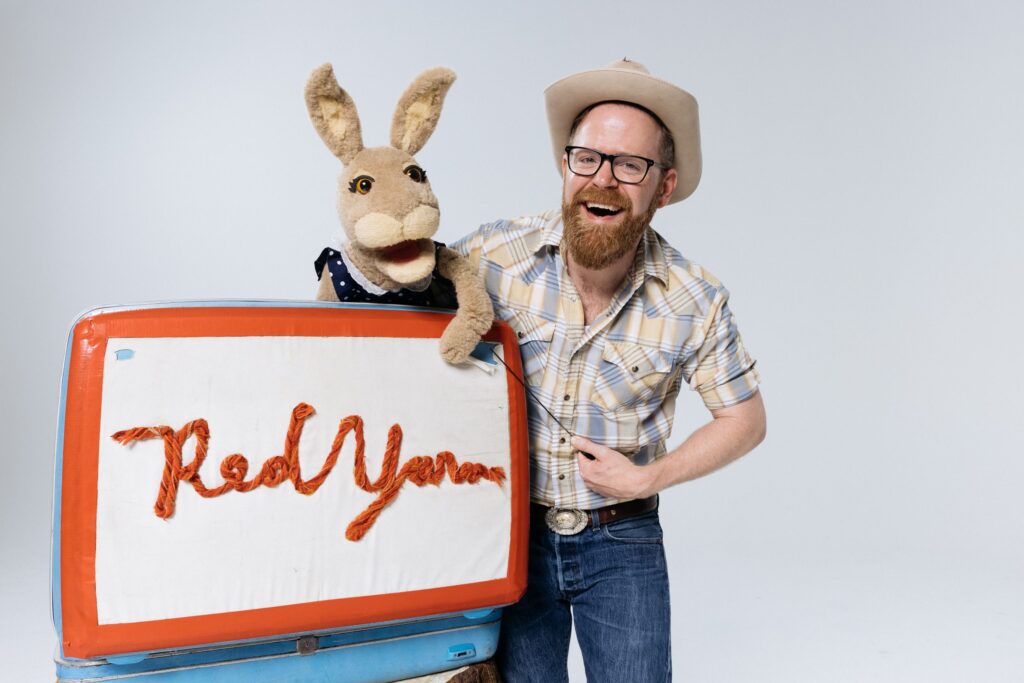 A bearded man in a cowboy hat smiles while holding a rabbit puppet. They both stand next to a prop that says "Red Yarn" in red yarn lettering. Red Yarn will perform as part of Wiggles and Giggles 2024