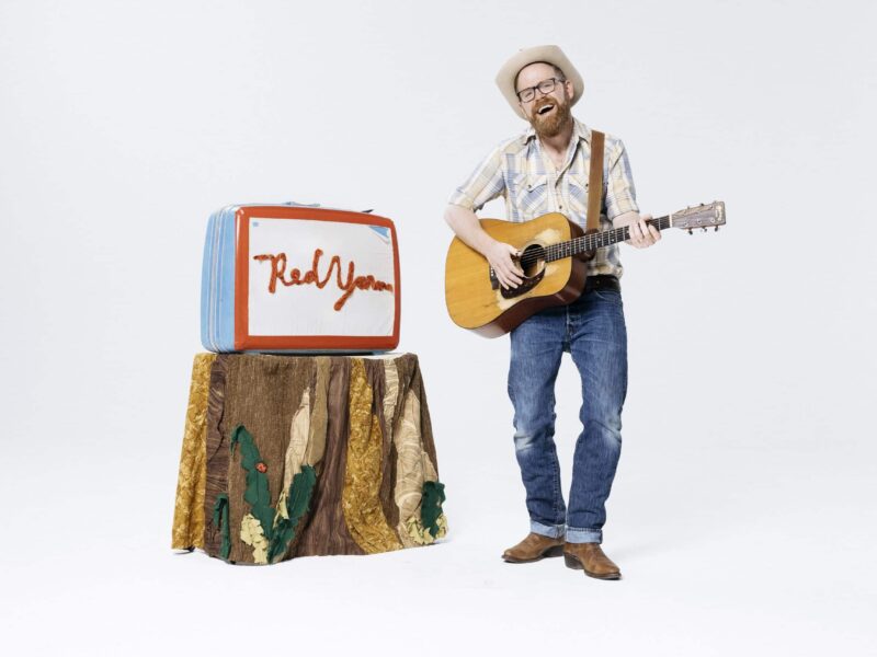 A bearded man in a cowboy hat smiles while playing guitar in front of a prop that says "Red Yarn" in red yarn lettering