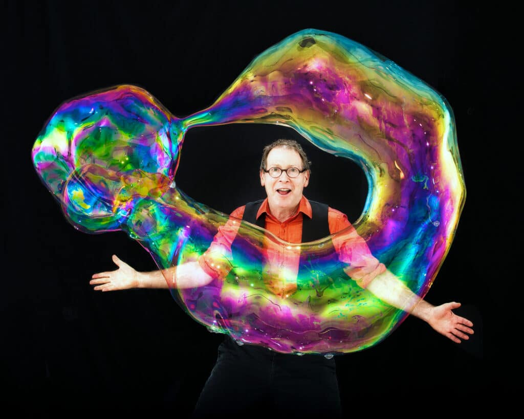 A man in glasses smiles behind a huge donut-shaped bubble in front of a black background. The Amazing Bubble Man will perform as part of Wiggles and Giggles 2024