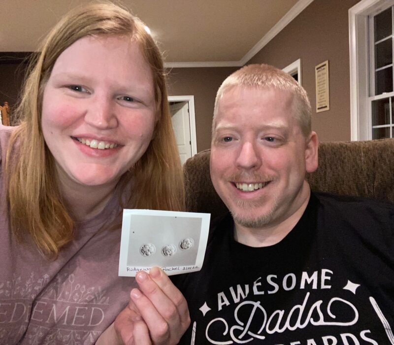 A man and woman smile while holding a photo of 3 embryos