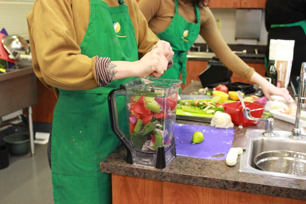 A person in a green apron prepares food next to a blender