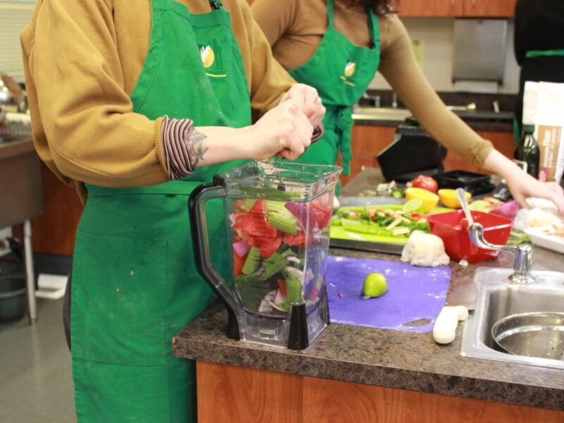 A person in a green apron prepares food next to a blender