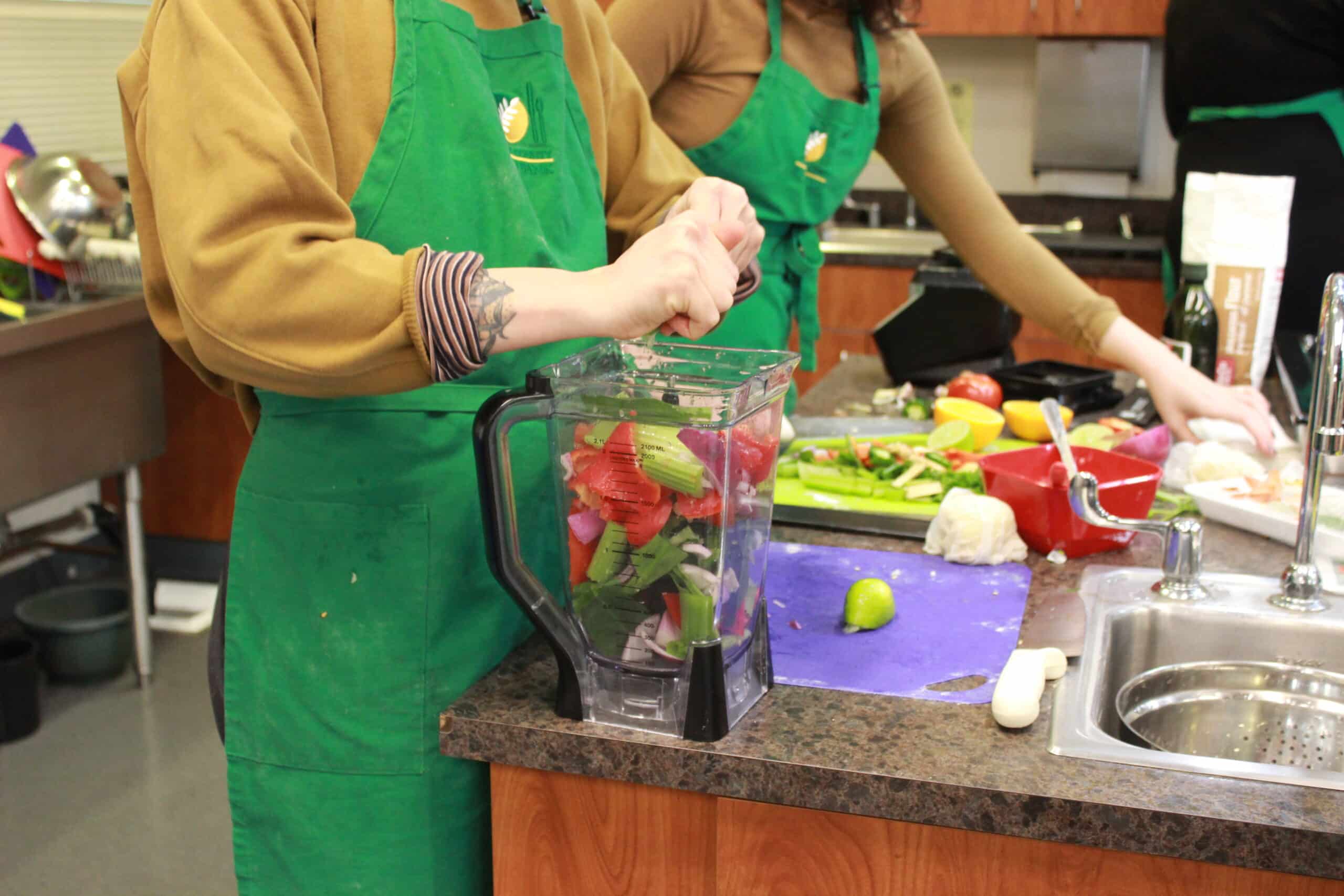 A person in a green apron prepares food next to a blender