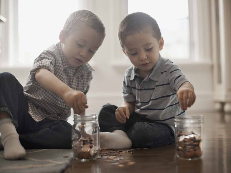 Two toddlers sit on the floor playing with coins and clear jars