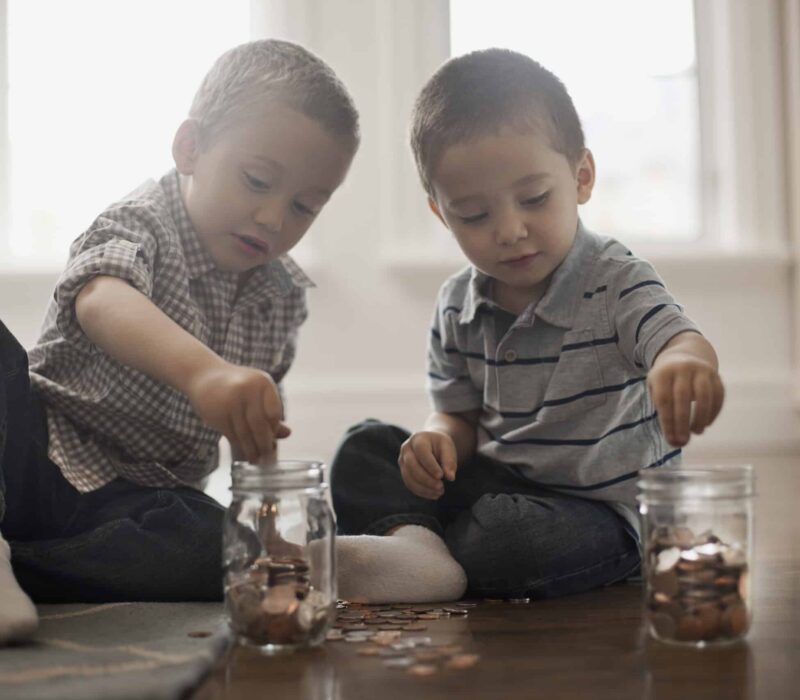 Two toddlers sit on the floor playing with coins and clear jars