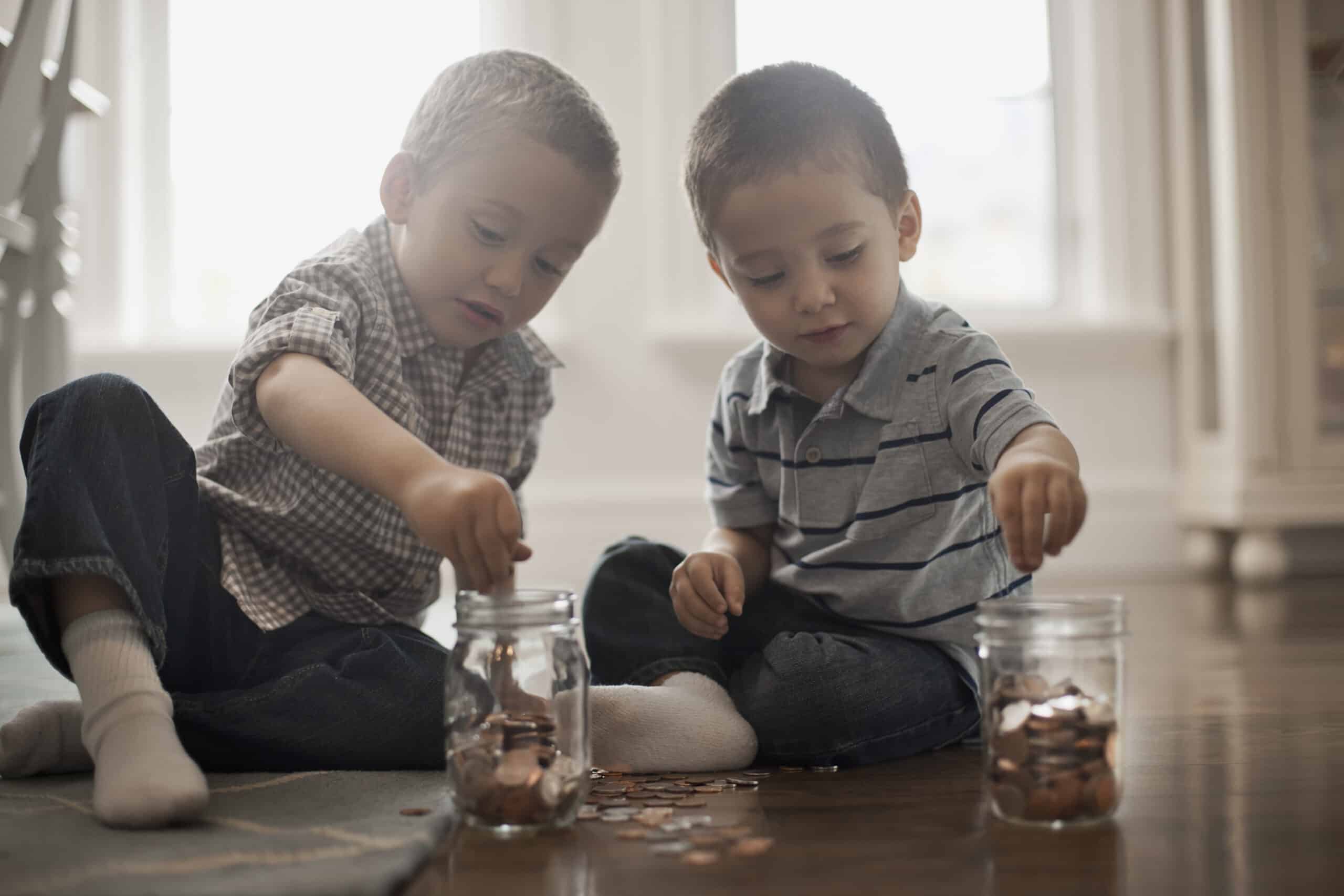 Two toddlers sit on the floor playing with coins and clear jars